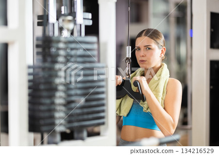 Girl in gym performs exercises for arm muscles using cable crossover machine and pull weighted rope 134216529