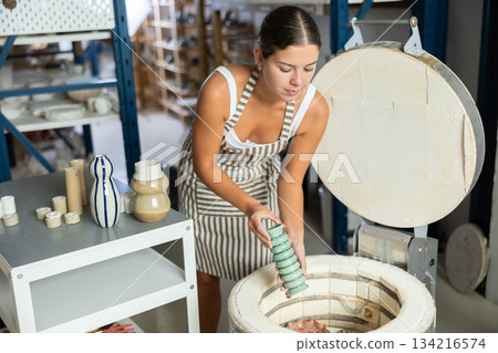 Young potter girl takes out finished ceramic dishes, various mugs and a vase from oven Young potter girl takes out finished ceramic dishes, various mugs and a vase from oven 134216574