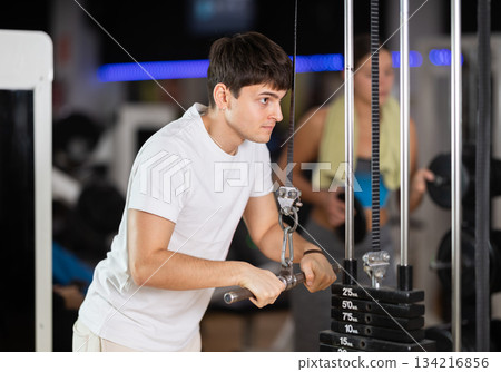 Guy in gym performs exercises for arm muscles using cable crossover machine and pull weighted rope Guy in gym performs exercises for arm muscles using cable crossover machine and pull weighted rope 134216856