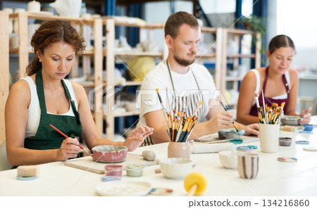 Three pottery masters work together on ceramic tableware, enjoying creative process as they paint mugs in workshop Three pottery masters work together on ceramic tableware, enjoying creative process as they paint mugs in workshop 134216860