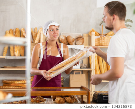 Saleswoman gives baguettes to young guy in bakery Saleswoman gives baguettes to young guy in bakery 134216878