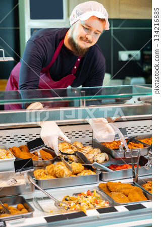 Salesman serving ready-to-eat dishes from deli counter in supermarket 134216885