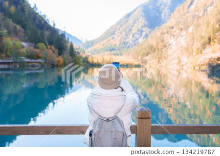 Happy woman traveler travel in jiuzhaigou national park in Sichuan, China. Tourist sightseeing and taking photo beautiful lake by mobile phone in Autumn season. Famous landmark near Chengdu, China 134219787