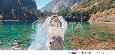 Happy woman traveler travel in jiuzhaigou national park in Sichuan, China. Tourist sightseeing Five Flower lake and snow mountain in Autumn season. Famous Landmark and Vacation near Chengdu, China 134219792