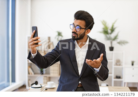 An Arab businessman in a suit gestures while on a video call using his smartphone An Arab businessman in a suit gestures while on a video call using his smartphone 134221179