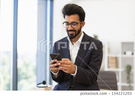An Indian businessman in a suit smiles while looking at his phone in an office setting 134221203
