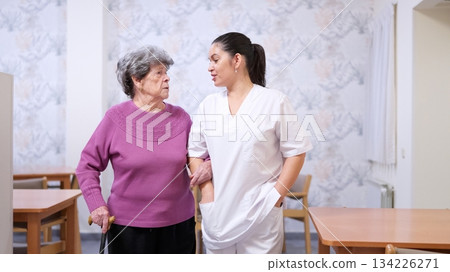 Caregiver assisting senior woman walking with cane in nursing home Caregiver assisting senior woman walking with cane in nursing home 134226271