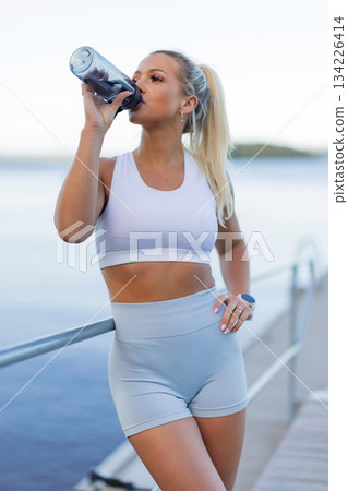 Active woman drinking water after outdoor workout beside a serene lake view Active woman drinking water after outdoor workout beside a serene lake view 134226414