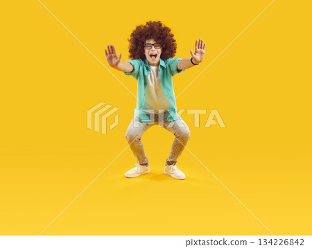 Happy funny little child in curly wig doing squats on orange color studio background Happy funny little child in curly wig doing squats on orange color studio background 134226842