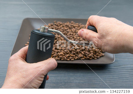 Hands operating a black manual coffee grinder over a tray of roasted coffee beans 134227159