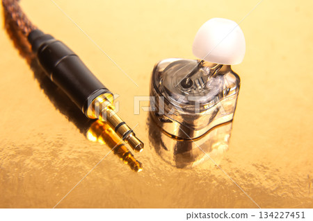 A single earbud, a gold-plated 3.5mm jack, and a black audio device on a reflective golden surface 134227451