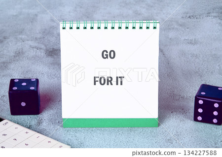 A notepad displaying GO FOR IT sits upright near dice and a calculator. The background has a mottled texture, suggesting a desk or workspace setting, beckoning chance and action 134227588