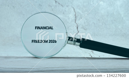 A magnifying glass highlights FINANCIAL FREEDOM 2026 on a neutral surface. It evokes ideas about achieving financial goals and planning for the future 134227600