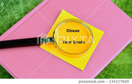 A magnifying glass highlights the phrase Chase Your Goals written on a vibrant yellow sticky note, set atop a cheerful pink book on a green surface 134227609