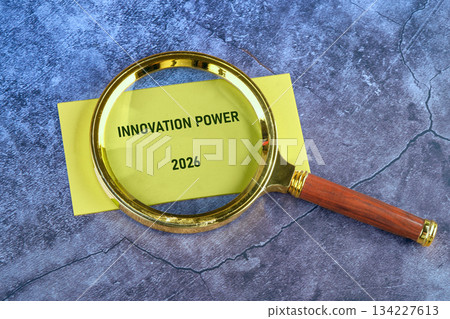A golden magnifying glass is placed on a yellow note, which displays the words Innovation Power and the year 2026. It rests upon a textured, marbled background 134227613