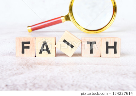 Wooden blocks spell out the word Faith, with the i block tilted. A golden magnifying glass lays above the word, on a white marble-like surface 134227614