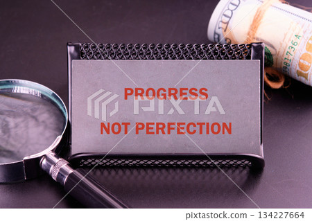 A sleek desk showcases a silver card holder displaying the powerful message of progress not perfection, accompanied by a magnifying glass and rolled cash, inspiring motivation and focus A sleek desk showcases a silver card holder displaying the powerful message of progress not perfection, accompanied by a magnifying glass and rolled cash, inspiring motivation and focus 134227664