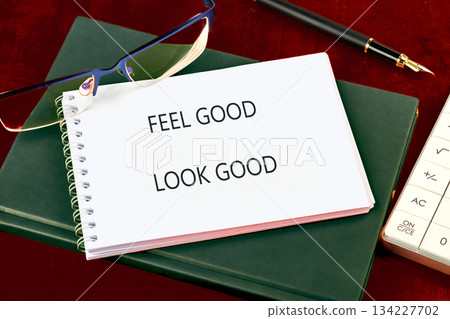 A spiral notepad shows the phrase FEEL GOOD LOOK GOOD. Nearby, glasses rest beside a black fountain pen, books, and a calculator on a dark burgundy surface A spiral notepad shows the phrase FEEL GOOD LOOK GOOD. Nearby, glasses rest beside a black fountain pen, books, and a calculator on a dark burgundy surface 134227702