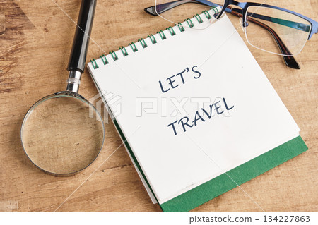 A notebook opened to a page that reads Lets Travel, accompanied by a magnifying glass and glasses. This scene inspires exploration and discovery of new places 134227863