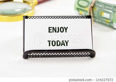 A clear sign displays the encouraging words Enjoy Today, suggesting a joyful approach to life. Nearby, a stack of money and a magnifying glass enhance the theme of appreciation 134227872
