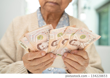 Asian elderly woman holding Thai banknotes money in hand. Poverty, saving problem in retirement. Asian elderly woman holding Thai banknotes money in hand. Poverty, saving problem in retirement. 134228653