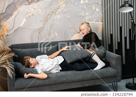 Children Couch Playing Pillow - Two kids playfully interact on a couch, one lying down while the other holds a pillow, capturing a lighthearted moment. 134228794