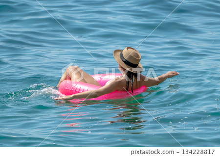 Woman Floating Sea. Back view of woman wearing straw hat on pink ring in ocean for summer relaxation. 134228817