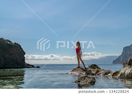 Summer, woman, sea. Young woman in red swimsuit stands on a rock in clear water, enjoying serene nature with copy space. 134228824
