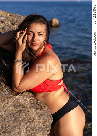 Woman bikini sea, young woman in red and black swimwear leaning on a rock by the ocean during summer vacation 134228860