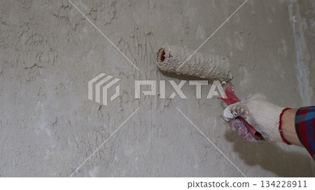 A hand in a fabric glove holds a roller against a wall covered with an uneven layer of plaster or mortar, applying putty and creating an uneven decorative coat on a wall indoors using a paint roller A hand in a fabric glove holds a roller against a wall covered with an uneven layer of plaster or mortar, applying putty and creating an uneven decorative coat on a wall indoors using a paint roller 134228911