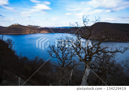 lake at the foot of a mountain surrounded by low mountains in autumn or winter surrounded by trees without leaves under a blue sky on the Edersee in Hesse Germany 134229419
