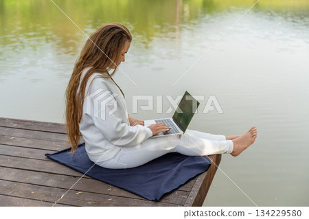 A woman is sitting on a dock with a laptop open in front of her. She is wearing white pants and a white sweater. The scene is peaceful and relaxing, with the water in the background. A woman is sitting on a dock with a laptop open in front of her. She is wearing white pants and a white sweater. The scene is peaceful and relaxing, with the water in the background. 134229580