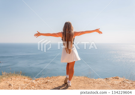 A woman stands on a rocky beach with her arms outstretched, looking out at the ocean. Concept of freedom and openness, as the woman seems to be embracing the beauty of nature. 134229604