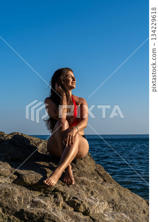 Woman relaxing on beach rocks, enjoying the sun by the sea under a clear blue sky with ample copy space Woman relaxing on beach rocks, enjoying the sun by the sea under a clear blue sky with ample copy space 134229618