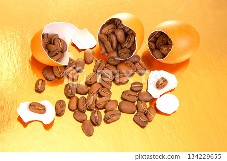 Creative still life of coffee beans emerging from a broken egg on a gold background Creative still life of coffee beans emerging from a broken egg on a gold background 134229655