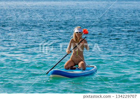 Paddleboarding, woman, ocean. Woman kneeling on stand-up paddleboard, paddling across blue ocean, enjoying summer recreation. 134229707