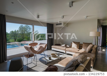 Modern luxury living room interior at sunset. Open-plan space featuring beige sofas, marble dining table, large glass windows with sun flare, and view of a patio. Stylish contemporary home design 134229753