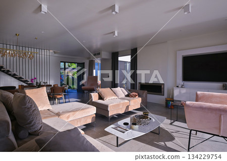 Modern luxury living room interior at sunset. Open-plan space featuring beige sofas, marble dining table, large glass windows with sun flare, and view of a patio. Stylish contemporary home design Modern luxury living room interior at sunset. Open-plan space featuring beige sofas, marble dining table, large glass windows with sun flare, and view of a patio. Stylish contemporary home design 134229754