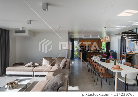 Modern luxury open-plan interior featuring a floating staircase, marble dining table, wooden kitchen, and beige sofa. Spacious contemporary home design with elegant lighting and gray tiled floors 134229763
