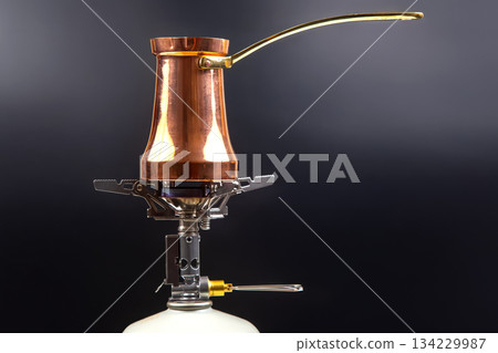 A shiny copper Turkish coffee pot with a brass handle sits atop a portable gas stove  134229987