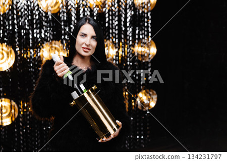 Charming woman with brunette long hair in trendy evening fur coat holding bucket with bottle with champagne on shine wall in studio 134231797