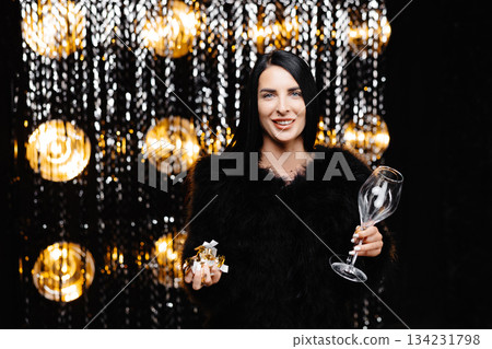 Happy woman with brunette long hair in trendy evening fur coat holding glass and confetti on shine wall in studio Happy woman with brunette long hair in trendy evening fur coat holding glass and confetti on shine wall in studio 134231798