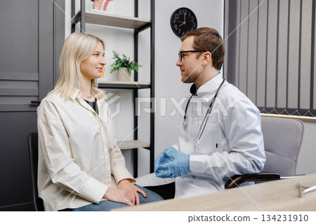 Vertical shot of young happy cheerful medical worker therapist in white doctor's robe having appointment consulting female patient in modern clinic hospital. Medical healthcare concept Vertical shot of young happy cheerful medical worker therapist in white doctor's robe having appointment consulting female patient in modern clinic hospital. Medical healthcare concept 134231910