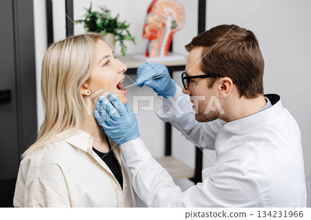 Doctor using inspection spatula to examine patient throat. ENT doctor doing throat exam of a woman. Patient opened her mouth to throat check-up Doctor using inspection spatula to examine patient throat. ENT doctor doing throat exam of a woman. Patient opened her mouth to throat check-up 134231966