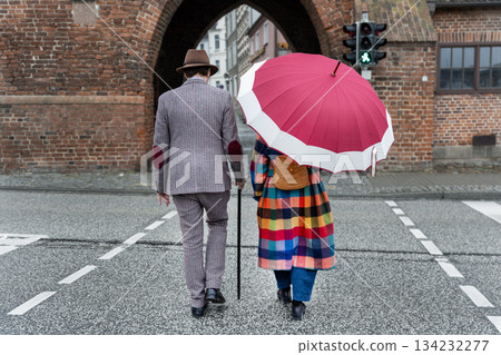 Stylish couple walks across street under bright umbrella near historic brick gate, wearing vintage clothing calm posture. Urban scene blends nostalgia color contrast quiet motion in European city Stylish couple walks across street under bright umbrella near historic brick gate, wearing vintage clothing calm posture. Urban scene blends nostalgia color contrast quiet motion in European city 134232277