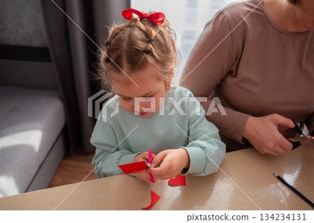 Top view of cute little girl cut out paper hearts sitting at a desk. Family is preparing for the celebration of Valentine's Day Top view of cute little girl cut out paper hearts sitting at a desk. Family is preparing for the celebration of Valentine's Day 134234131