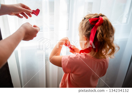 Parent and her daughter decorate the curtains on the window with red paper hearts. Rear view. A celebration for Valentine's Day and Mother's Day 134234132
