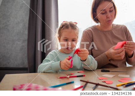 Mother and her little daughter cut out paper hearts sitting at a desk. Family is preparing for the celebration of Valentine's Day Mother and her little daughter cut out paper hearts sitting at a desk. Family is preparing for the celebration of Valentine's Day 134234136