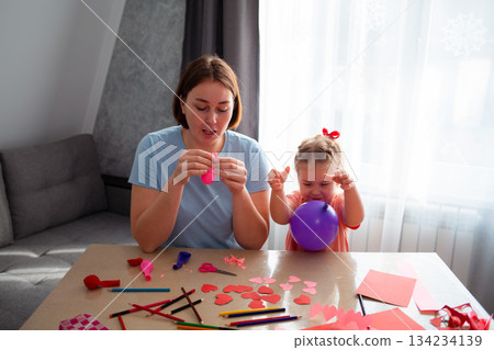 Mid shot of young Caucasian mother and her cute little daughter inflate balloons and make decorations for the holiday. Family sits at the table and makes festive decorations Mid shot of young Caucasian mother and her cute little daughter inflate balloons and make decorations for the holiday. Family sits at the table and makes festive decorations 134234139