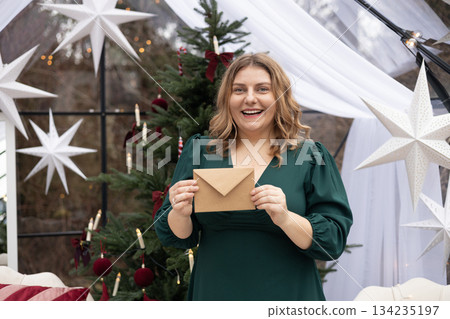 Young 30s woman getting Christmas greeting card from her beloved on background of xmas tree at cozy living room with festive interior. Surprised lady pleasure reading letter at home. Young 30s woman getting Christmas greeting card from her beloved on background of xmas tree at cozy living room with festive interior. Surprised lady pleasure reading letter at home. 134235197